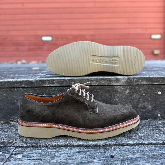 The ALDEN SHOES D5426 - Loden Suede Plain Toe Blucher is a dark brown suede oxford with beige laces and a chunky tan wedge sole, shown on a weathered wooden surface with a red building in the background.