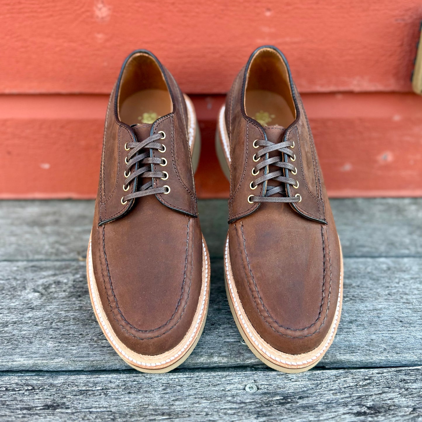 A pair of ALDEN SHOES 73980 - Tobacco Chamois Ranger Mocc with Wedge lace-ups sits on a weathered wooden surface before a red wall.