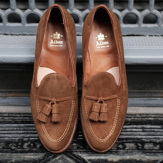 A pair of Alden Madison 3403 Tassel Loafers in snuff suede, featuring a leather sole and Aberdeen Last, sits side by side on a textured dark surface before a decorative metal grate.