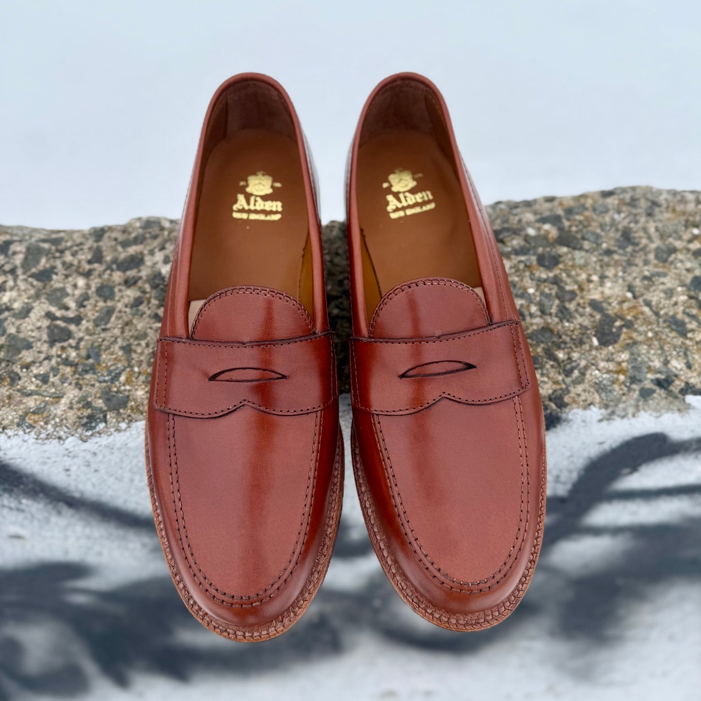 A pair of tan Michigan calf 9823 Penny Loafers by Alden Madison, with oak leather soles and the gold Alden logo inside, displayed side by side on a textured stone surface.