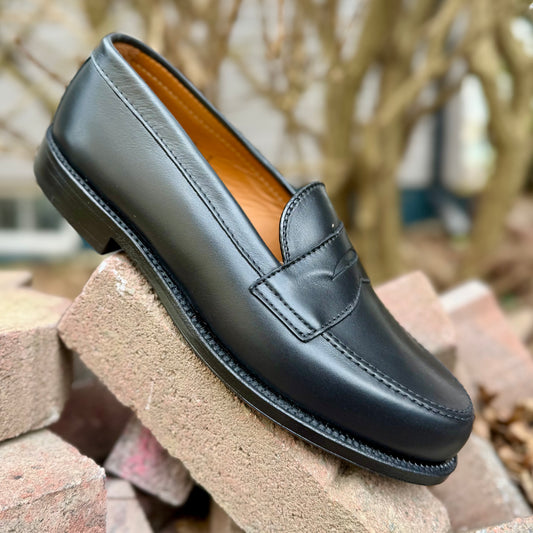 The ALDEN SHOES 9827 - Black Calf Leisure Penny Loafer, featuring visible stitching and a single leather sole, rests angled on light brown bricks. Bare tree branches in the blurred background highlight its classic Van Last design.