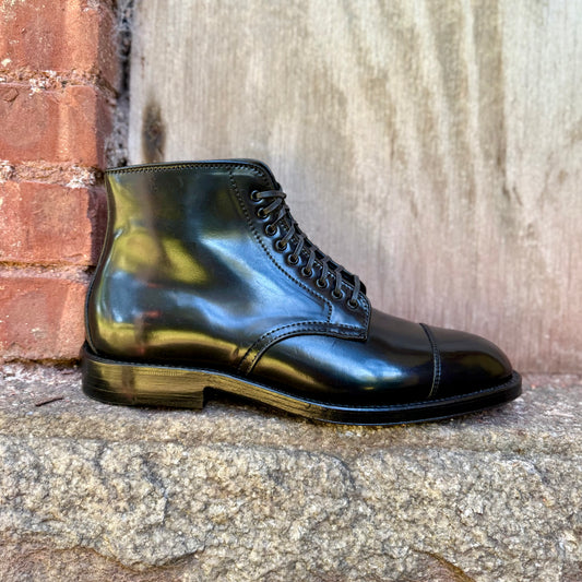 The Alden Madison 4065 Cap Toe Boot in Black Shell Cordovan, crafted on the Grant Last with a Double Oak Leather Sole, is showcased on a stone surface against a brick wall and wooden background.
