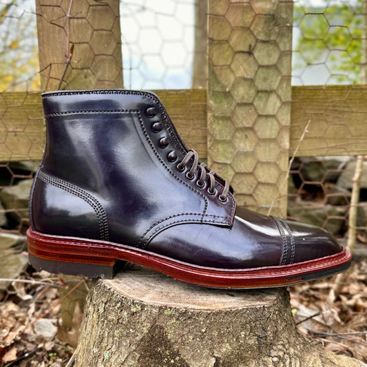 Alden Madisons D2858HC - 2/2 Cap Boot w Commando in Color 8 Shell sits on a stump. With a brown sole, its polished black leather and refined charm stand out against the fence and scattered leaves in the background.