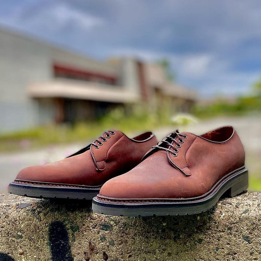 A pair of Alden Madisons 9431S - Plain Toe Blucher in Oiled Nubuck sits on a stone surface outdoors. The slightly blurred background reveals structures and greenery under a partly cloudy sky, highlighting every rugged yet elegant step in Tobacco Oiled Nubuck.