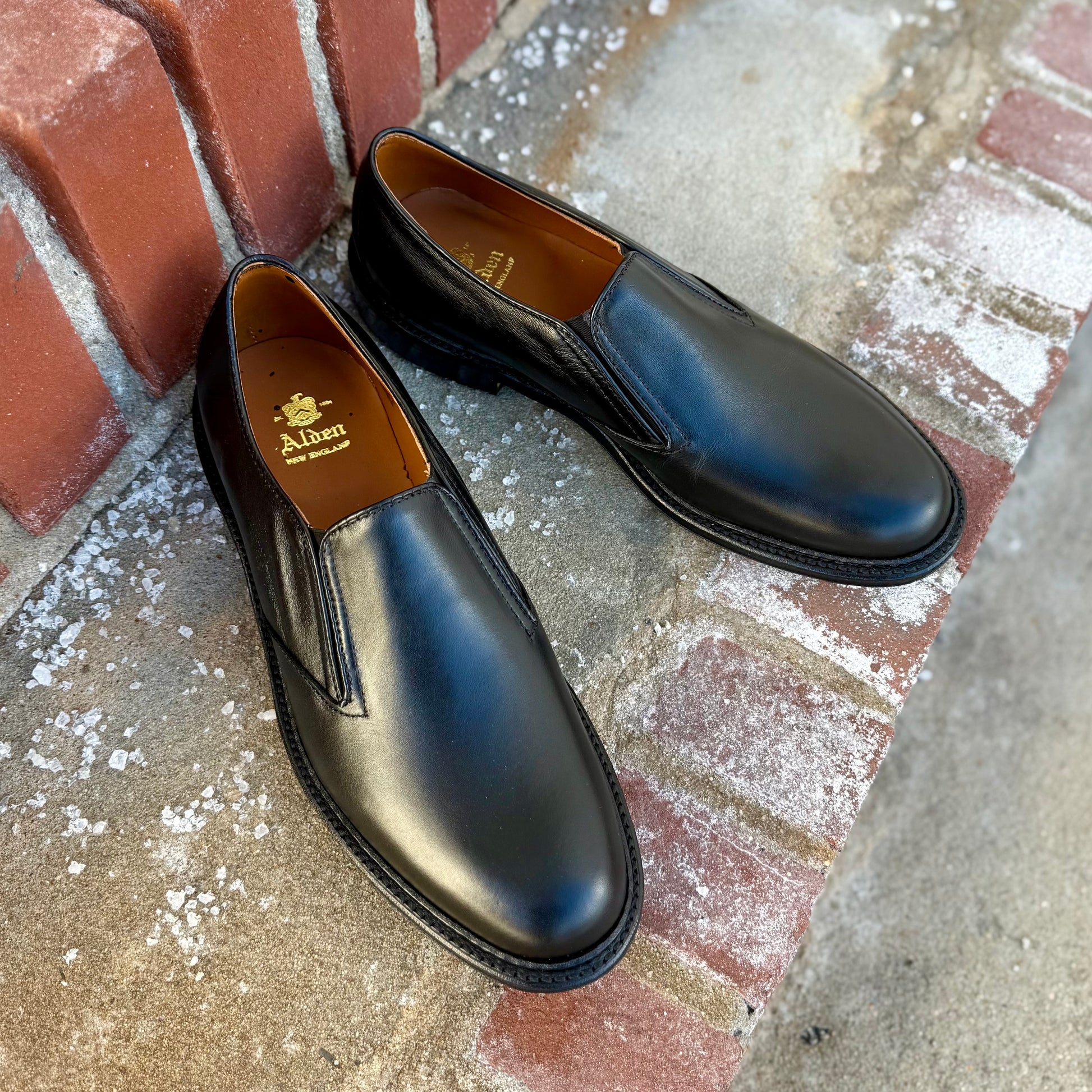 A pair of clean, shiny black ALDEN SHOES D2406L "Double Gore" Slip On Rubber Sole dress shoes displayed on a red brick and concrete surface, with visible Alden branding inside.
