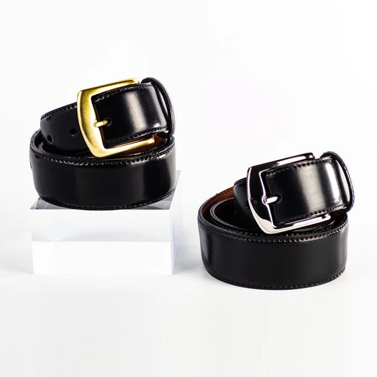 Two Alden Madison 38mm Black Shell Cordovan Belts—one with a nickel buckle, one with a brass buckle—are displayed on a white background. The brass-buckle belt is elevated on a clear acrylic block. Made in USA.
