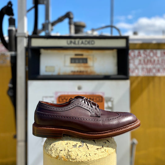 The Alden Madison 976 Long Wing Blucher in polished brown calfskin, featuring brogue detailing and a double waterlock sole, is displayed on a concrete pillar. An old-fashioned UNLEADED gas pump stands under a partly cloudy sky in the background.