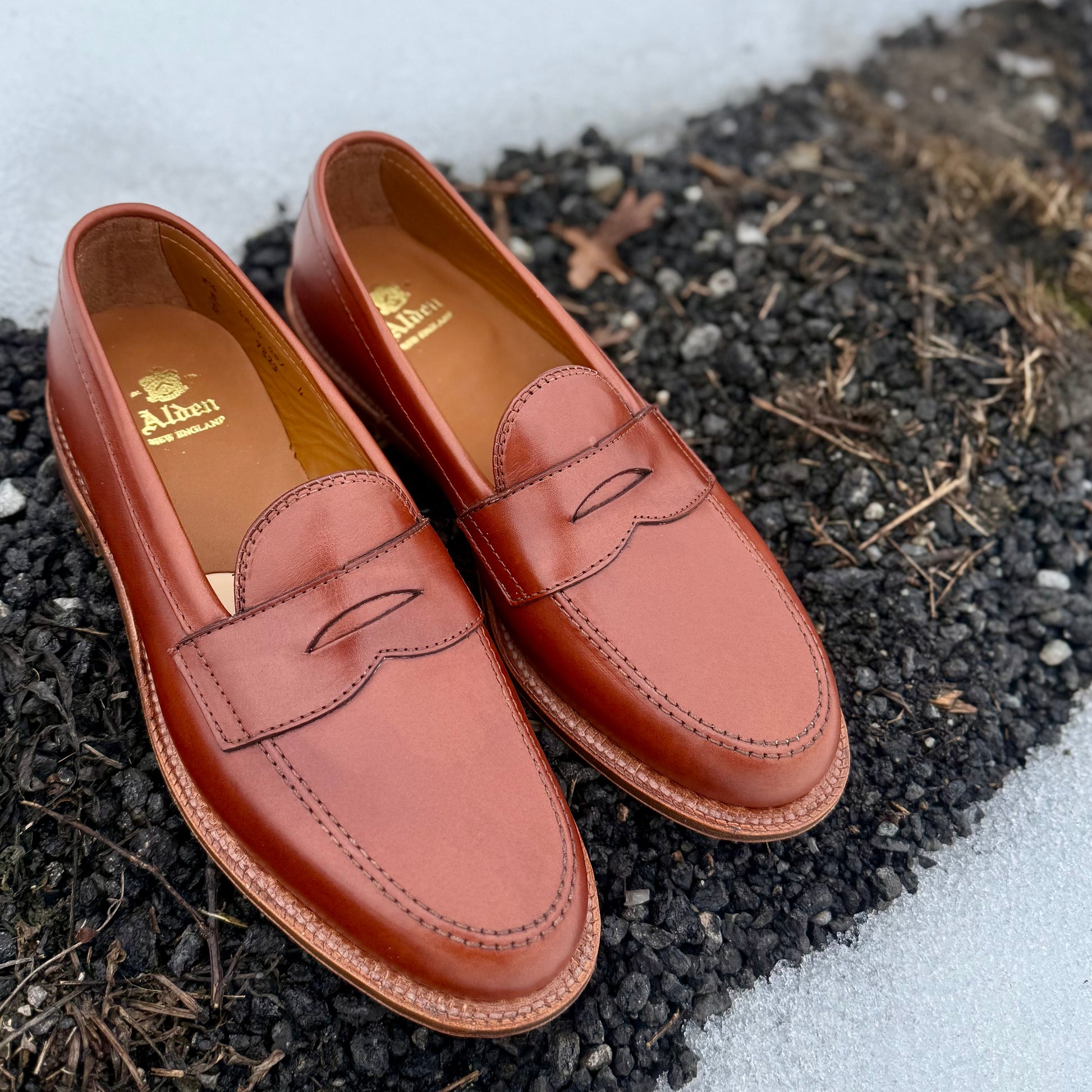 A pair of Alden Madison 9823 - Penny Loafer in Tan Michigan Calf with Oak Leather Sole is set outdoors on dark gravel and dirt, with patches of snow visible in the background.