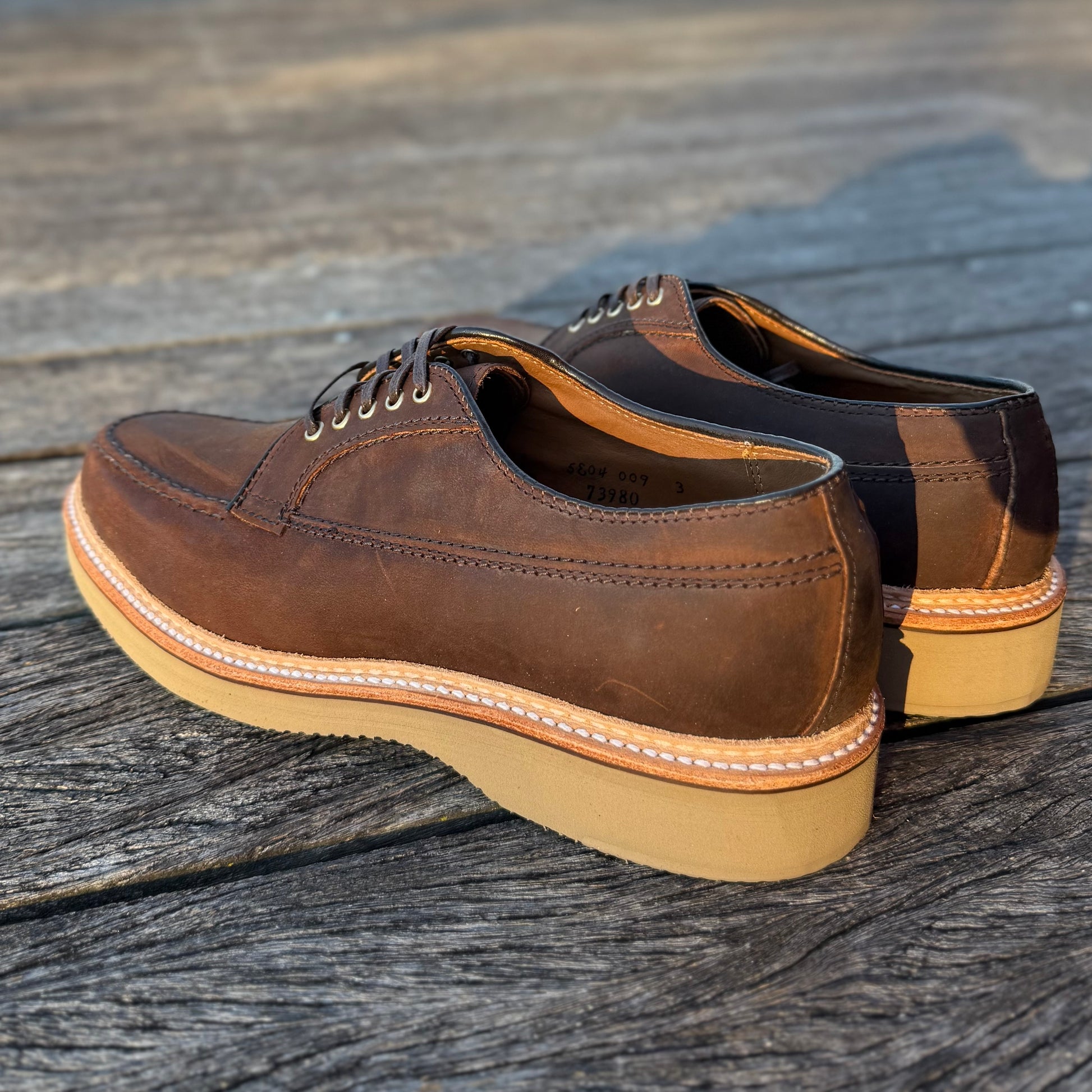 A pair of ALDEN SHOES 73980 - Tobacco Chamois Ranger Mocc with Wedge rests on a weathered wooden surface in sunlight, highlighting the brown leather, light tan wedge soles, and white stitching.