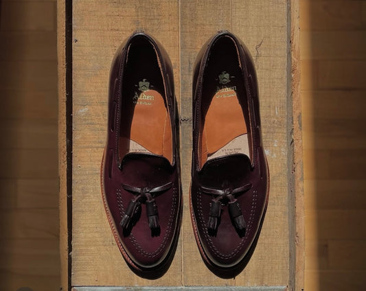 Alden Madison’s D7103 - Antique Tassel Loafer in Color 8 Shell Cordovan, crafted on the Aberdeen Last with an Oak Leather Outsole, is displayed side by side on a wooden surface as sunlight casts shadows around them.