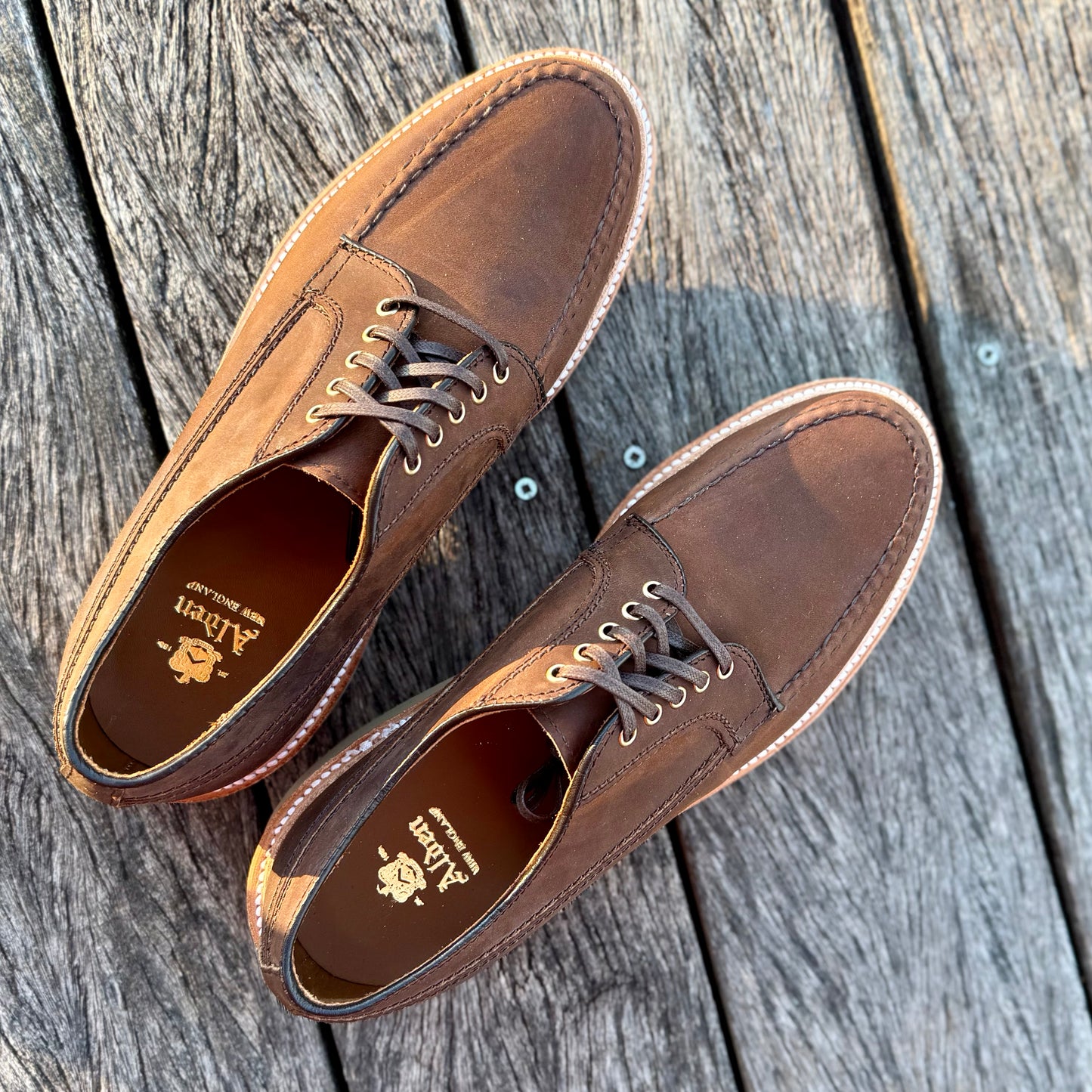 A pair of 73980 - Tobacco Chamois Ranger Mocc with Wedge from ALDEN SHOES, featuring lace-up design, stitching details, and a comfy wedge sole, shown on a weathered wooden surface with the brand visible inside.