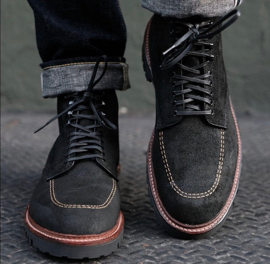 A close-up of a person wearing cuffed jeans and Alden Madison’s D9916H - Indy Boot in Earth Reverse Chamois, featuring black suede, contrast stitching, brown soles, and a Vibram Heavy Lug Outsole on a textured metal surface.
