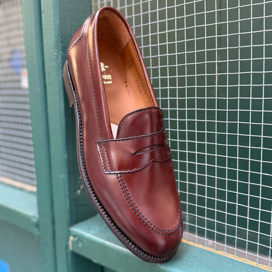 A single Alden Madison D5214 - Brown Calf Flex Penny loafer, crafted from luxurious Brown Vegano Calf leather with fine stitching detail, rests against a teal wooden surface and wire mesh background.