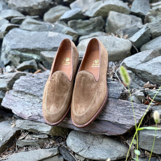 The Alden Madison D2101 - Vanderbilt Slip On in snuff suede, featuring stitched detailing and a leather sole, is showcased on flat rocks outdoors amid larger gray stones and grass.