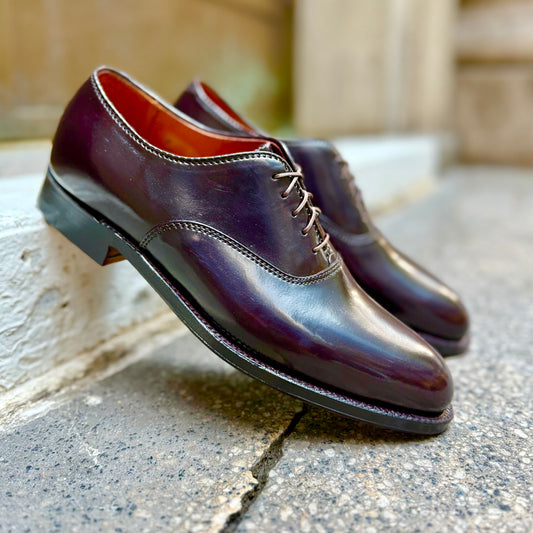 A pair of Alden Madison 934 - Plain Toe Bal dress shoes in Color 8 Shell Cordovan with laces rests on a white ledge, set against a textured outdoor surface.