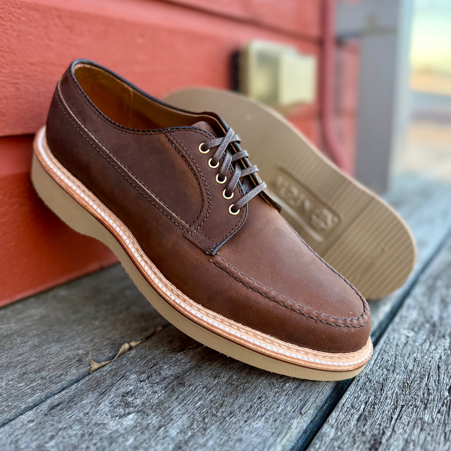 A pair of ALDEN SHOES 73980 - Tobacco Chamois Ranger Mocc with Wedge is displayed on a wooden surface before a red wall, with one shoe upright and the other leaning against it.