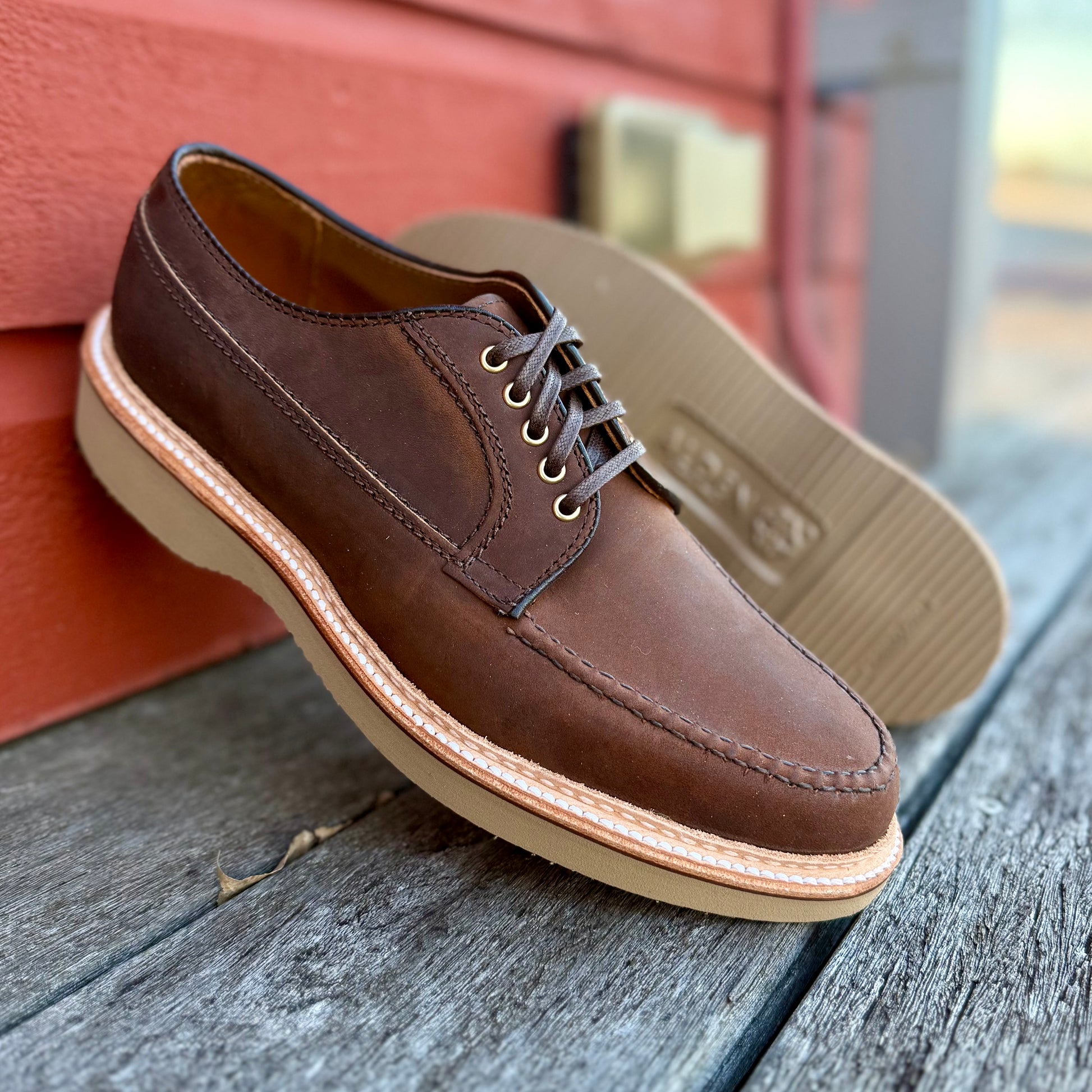 A pair of ALDEN SHOES 73980 - Tobacco Chamois Ranger Mocc with Wedge is displayed on a wooden surface before a red wall, with one shoe upright and the other leaning against it.