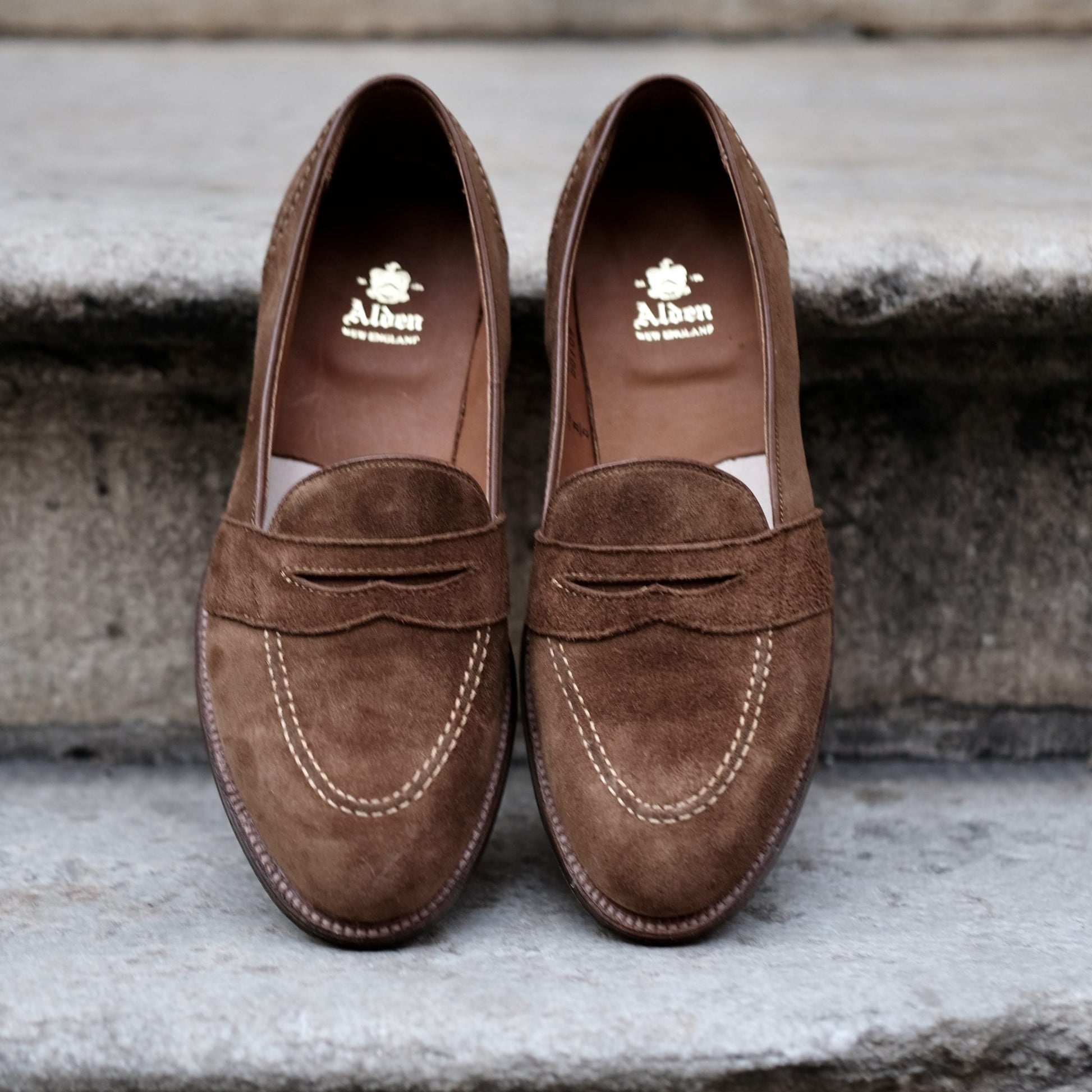 Alden Madison’s D0109 - Full Strap Loafer in snuff suede, featuring white stitching and leather outsoles, displayed side by side on stone steps. Gold Alden logo details the insoles.