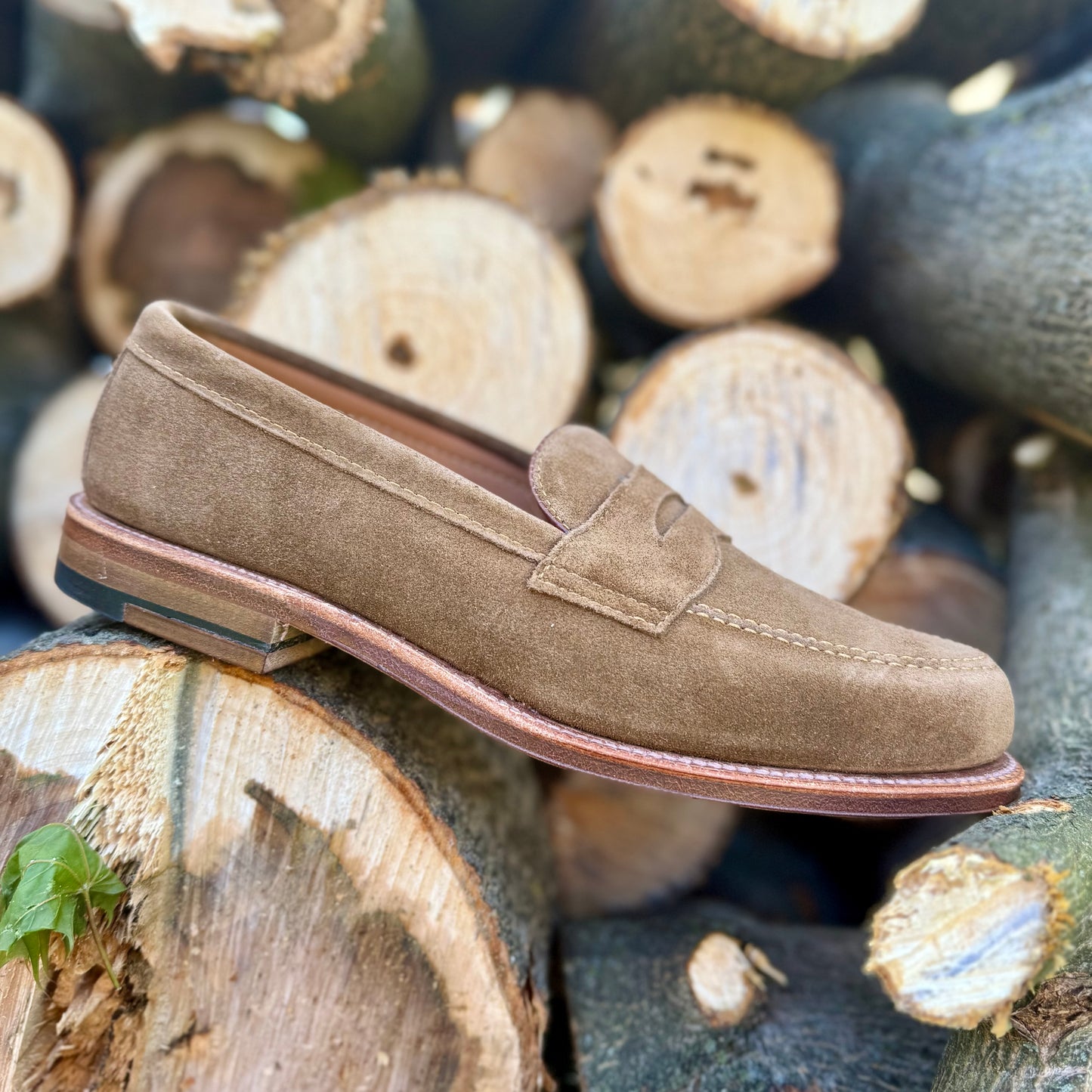 The Alden Madison 6173F - Penny Loafer in Snuff Suede is shown against stacked logs, showcasing its rich texture and Flex Leather Sole craftsmanship.