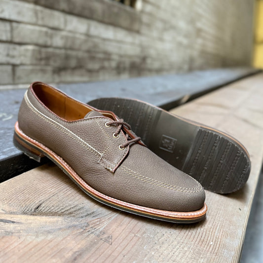 Alden Madison’s 7351 - Ivy Blucher in Clay Nubuck sits on a wooden surface—one upright, the other sideways to reveal the sole—against a blurred brick wall, showcasing classic brown lace-up style and premium craftsmanship.