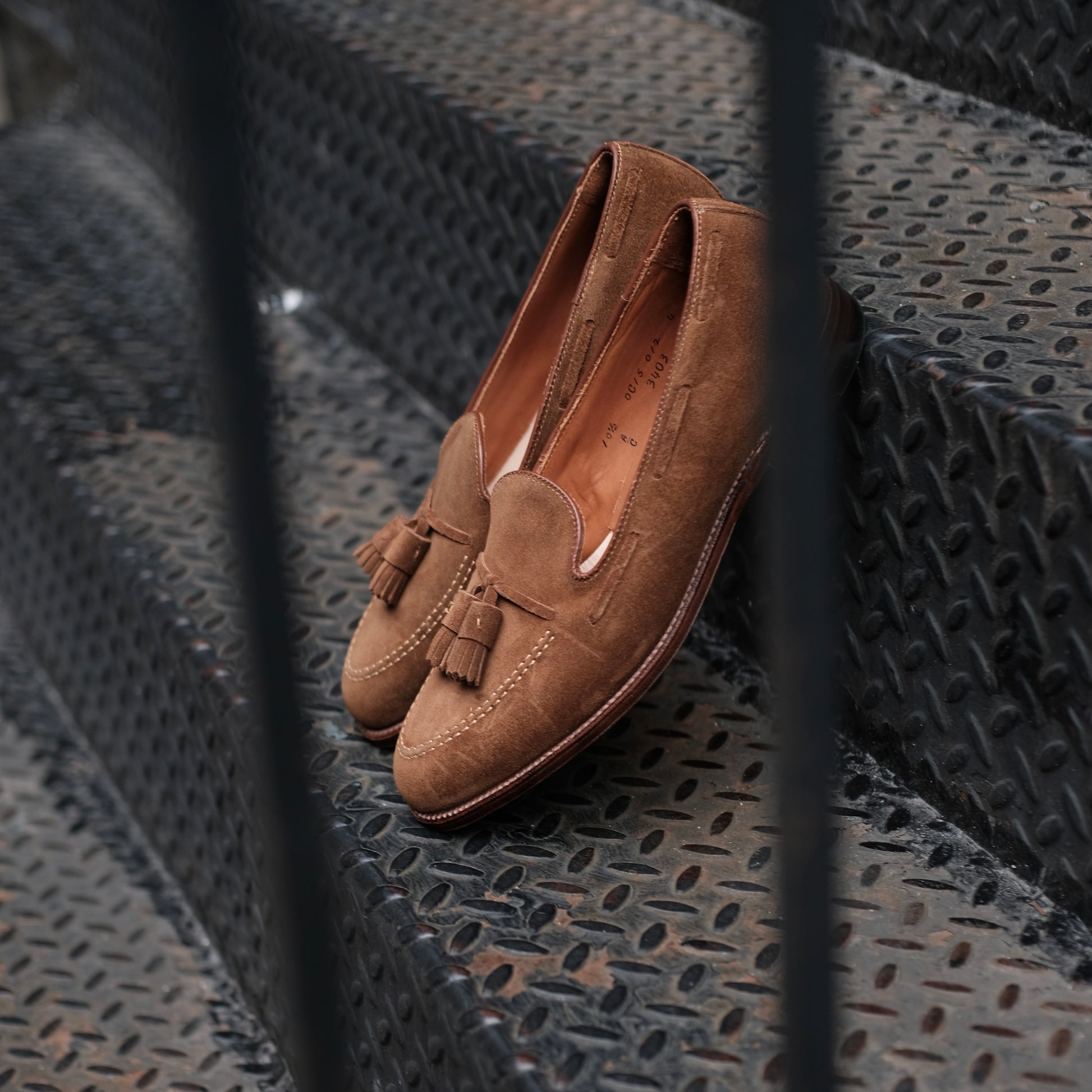 The Alden Madison 3403 Tassel Loafer in snuff suede, featuring a leather sole and crafted on the Aberdeen Last, is displayed on a black metal staircase behind vertical railings.