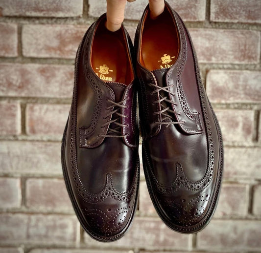 A hand holds up a pair of Alden Madison 975 - Long Wing Blucher shoes in Color 8 Shell Cordovan, featuring brogue detailing and a Double Oak Leather Sole, in front of a light-colored brick wall.