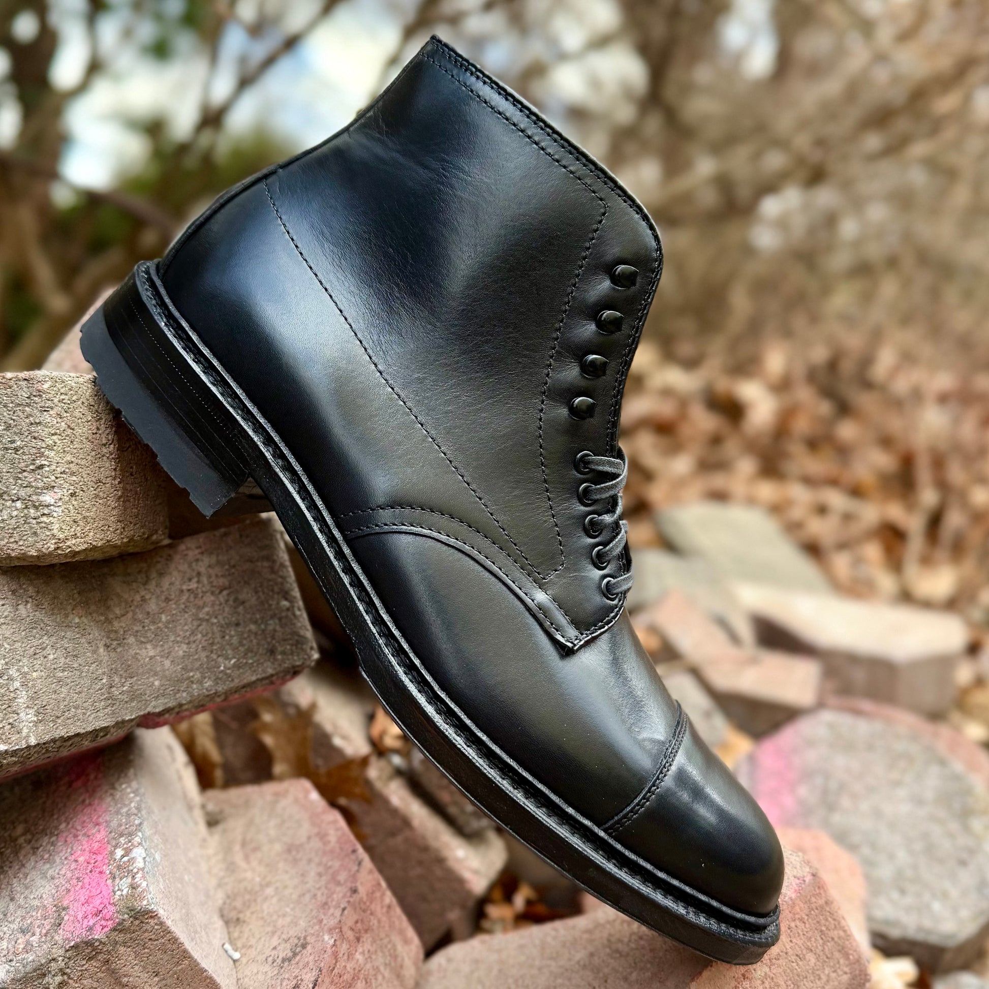 The Alden Madison 40797HC Black Cap Toe Boot with Commando Sole, styled on a Barrie last, is displayed outdoors on stacked bricks with a blurred background of dry leaves and trees.