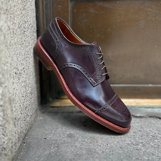 The Alden Madison 21522 - Medallion Cap Toe Blucher Color 8 shoe, featuring a polished reddish-brown leather finish with decorative perforations and a double leather sole, is propped against a concrete wall near a metal door.