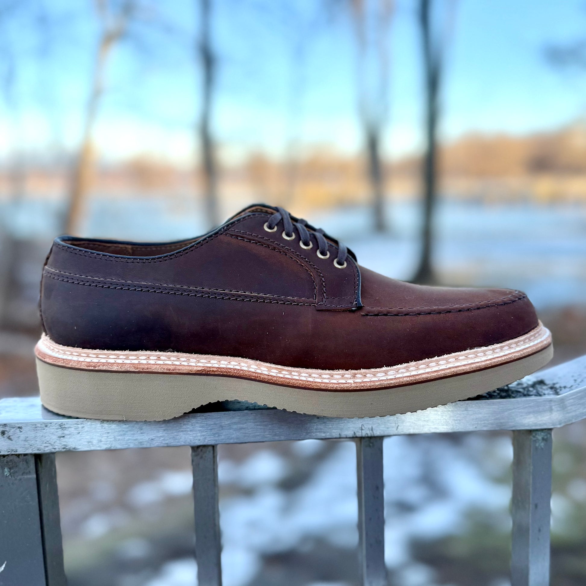 The ALDEN SHOES 73980 - Tobacco Chamois Ranger Mocc with Wedge is shown on a metal railing outdoors, featuring brown leather, lace-up design, and a light tan wedge sole, set against a blurred backdrop of trees and snow.