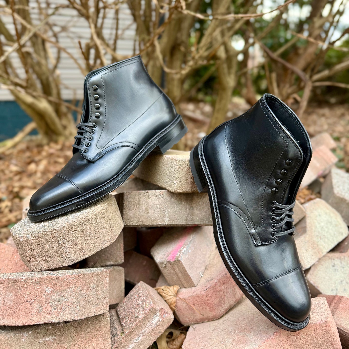 The Alden Madison 40797HC Black Cap Toe Boot with Commando Sole is shown outdoors atop bricks, with leafless branches and a building softly blurred in the background.