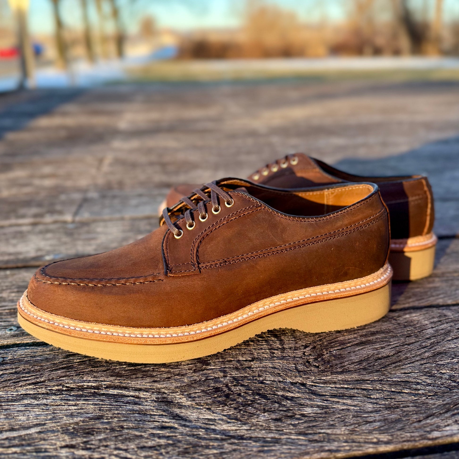 A pair of ALDEN SHOES 73980 - Tobacco Chamois Ranger Mocc with Wedge sits on a wooden surface outdoors, blurred trees and grass in the background.