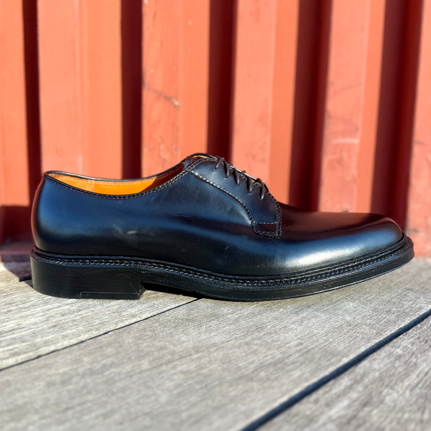 The ALDEN SHOES 9909 Black Calfskin Plain Toe Blucher with laces rests on a wooden surface in bright sunlight, set against a red paneled wall.