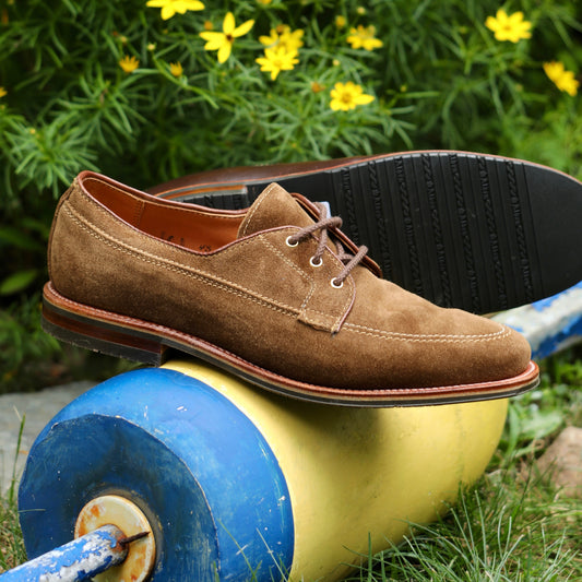 The ALDEN SHOES 735L - Ivy Blucher in Snuff Suede is shown outdoors, with one shoe upright and one on its side atop a yellow and blue buoy, set against green foliage and yellow flowers.