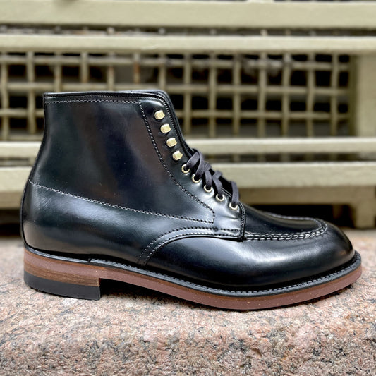 The Alden Madison D1908H Copake Indy Boot in Black Shell, featuring gold eyelets, hand stitching, and a brown sole, is displayed on a stone surface with a metal grate backdrop.