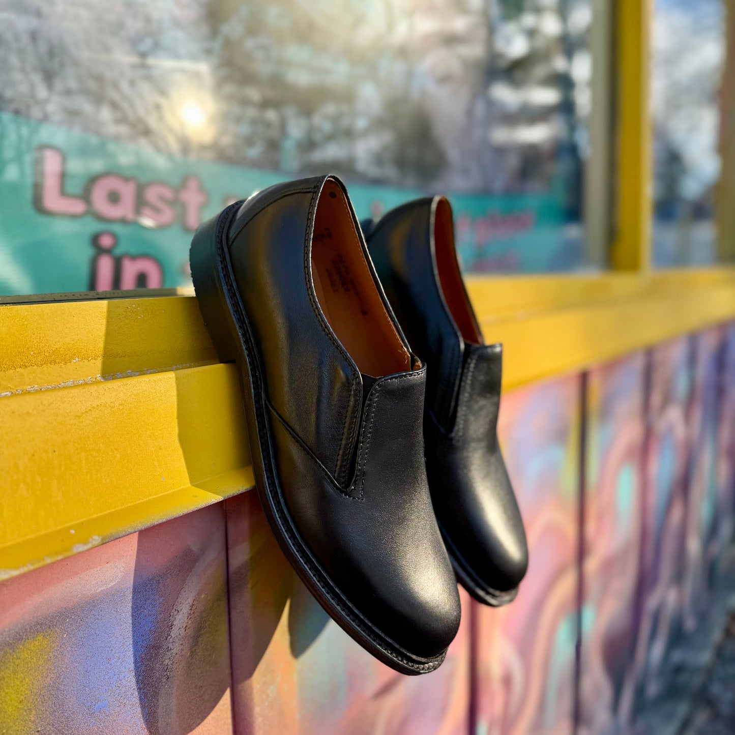 A pair of ALDEN SHOES D2406L "Double Gore" black calfskin slip-on dress shoes with rubber soles sits on a yellow ledge before a colorful graffiti wall, sunlight casting shadows and a blurred sign in the background.