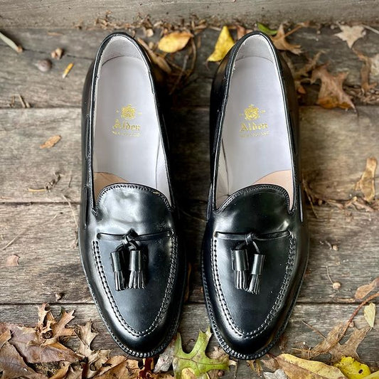 The 664 Tassel Loafers in Black Shell Cordovan by Alden Madison rest elegantly on a wooden surface, surrounded by scattered autumn leaves, with the brands name gracing the insoles, showcasing craftsmanship at its finest.