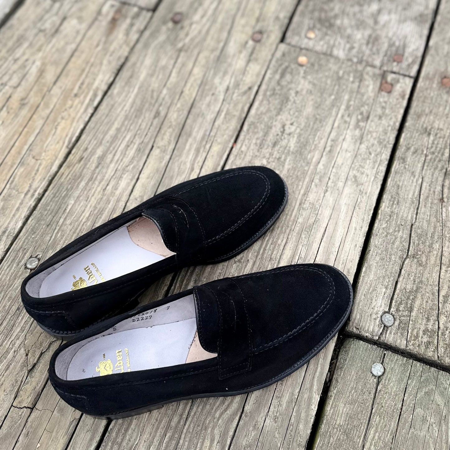 The Alden Madison D2229 “Stuyvesant” penny loafers in black suede, with white insoles and classic leather soles, are displayed side by side and slightly angled on a weathered wooden deck.