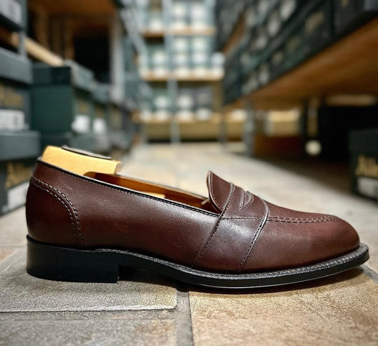 Alden Madisons 686 - Full Strap Loafer in Brown Calfskin features a black sole and is crafted on the classic Aberdeen Last, pictured side view on a tiled floor with shelves of shoe boxes in the background.