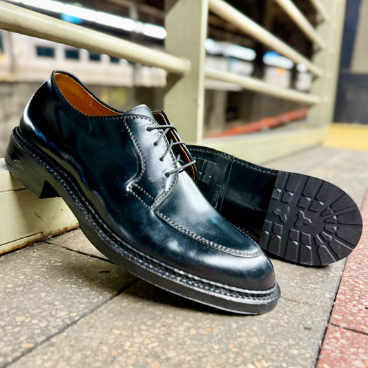 A pair of ALDEN SHOES D4604C - Moc Toe “Borough” Bluchers in Black Shell Cordovan, one upright and one on its side displaying the sole, are shown on a tiled floor at a train or subway station.