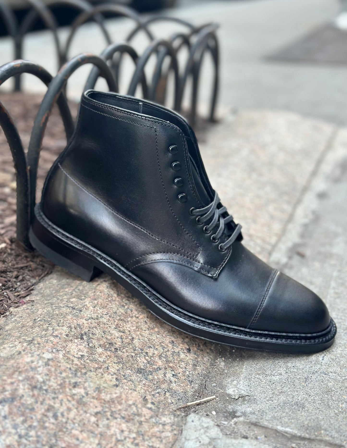 The Alden Madison 40797HC Black Cap Toe Boot with Commando Sole sits on a stone curb by a metal fence, with the sidewalk and street softly blurred in the background.