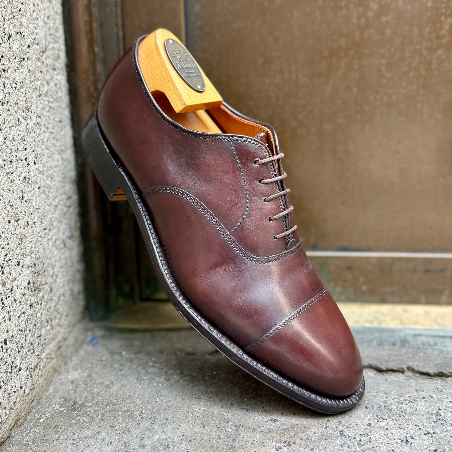 The Alden Madison 920 - Cap Toe Bal in Brown Calfskin, shown with a wooden shoe tree and single oak leather sole on the Hampton last, leans against a concrete and metal surface.