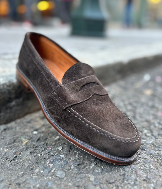 Close-up of the Alden Madison 6248F Unlined Leisure Hand Sewn in Dark Chocolate Suede. The penny loafer features classic stitching, detailing, a slight heel, and a visible Flex Leather Sole edge against a blurred concrete background to emphasize its texture and craftsmanship.