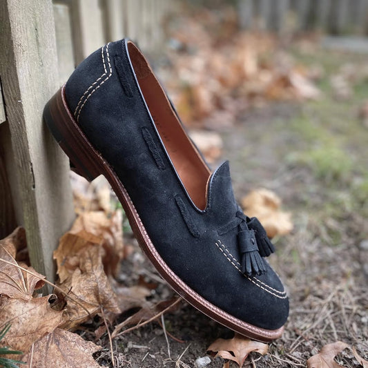 The Alden Madison D0106 - Black Suede Tassel Loafer, featuring white stitching and crafted on the Aberdeen Last, rests by a wooden fence among dry brown leaves and grass.