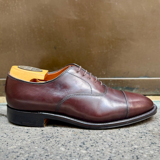 The Alden Madison 920 Cap Toe Bal in brown calfskin, with a single oak leather sole and wooden shoe tree inserted, is displayed on a gray textured surface against a metallic background.