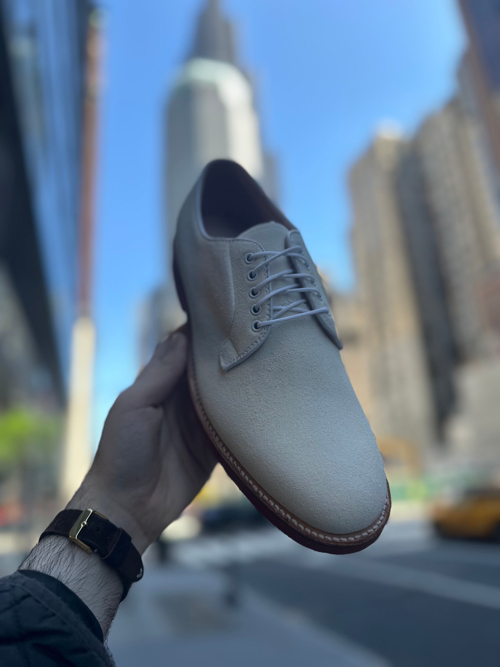 A hand holds up the Alden Madison 9448 - White Buck in Ivory Suede with a Microcell Sole against a city street and skyscrapers, while wearing a black watch.