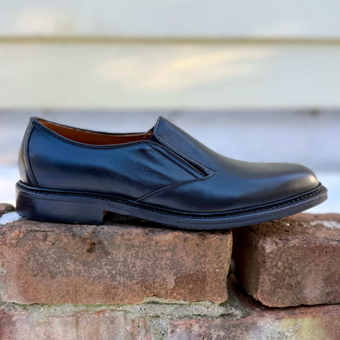 The ALDEN SHOES D2406L "Double Gore" Slip On, a black calfskin ladies’ shoe with a low heel and rubber sole, is shown on a brick surface with a blurred background.