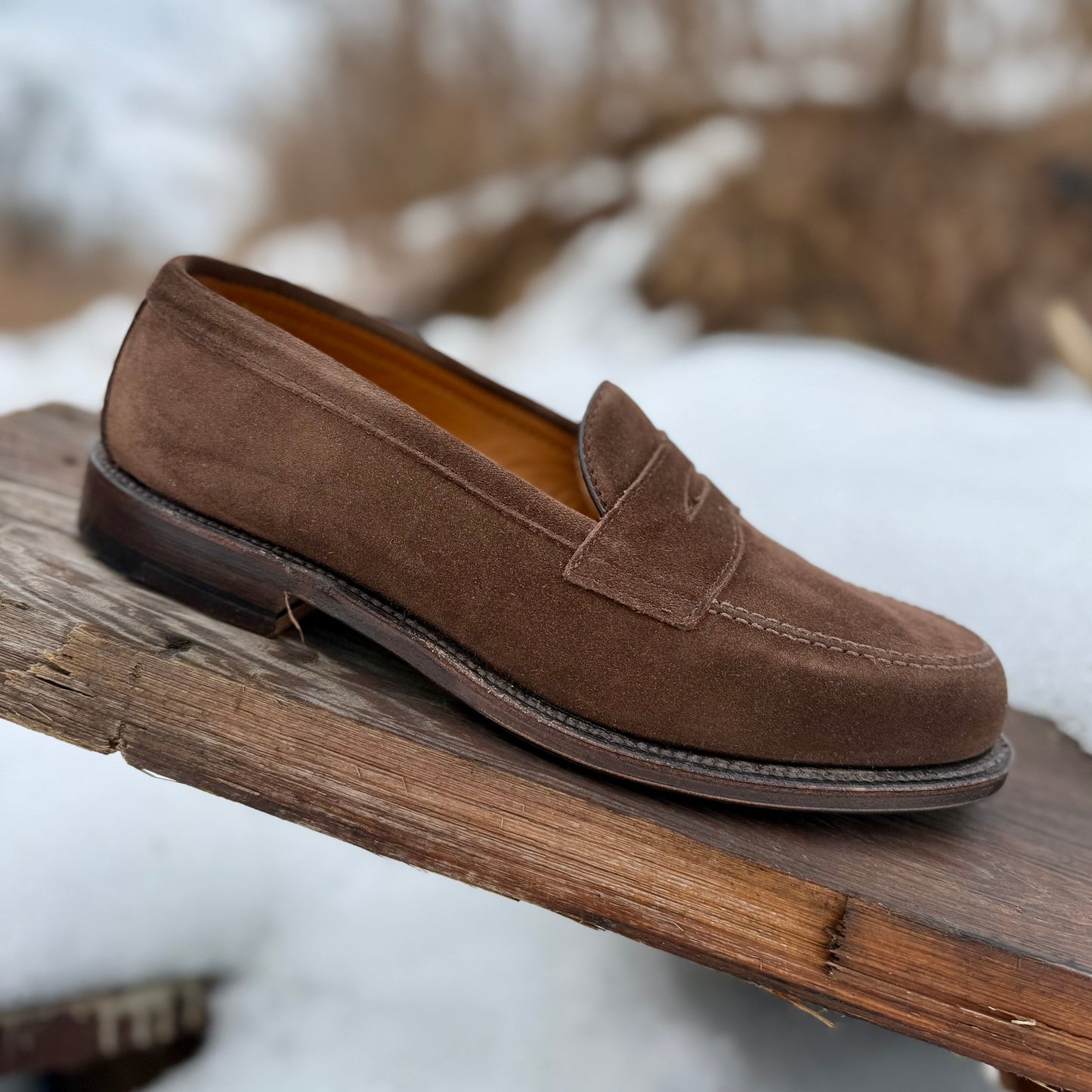 The Alden Madison 6175F Penny Loafer in Humus Suede, featuring a dark brown Flex Oiled Leather Sole, rests on a weathered wooden plank outdoors with snow and blurred natural scenery in the background.