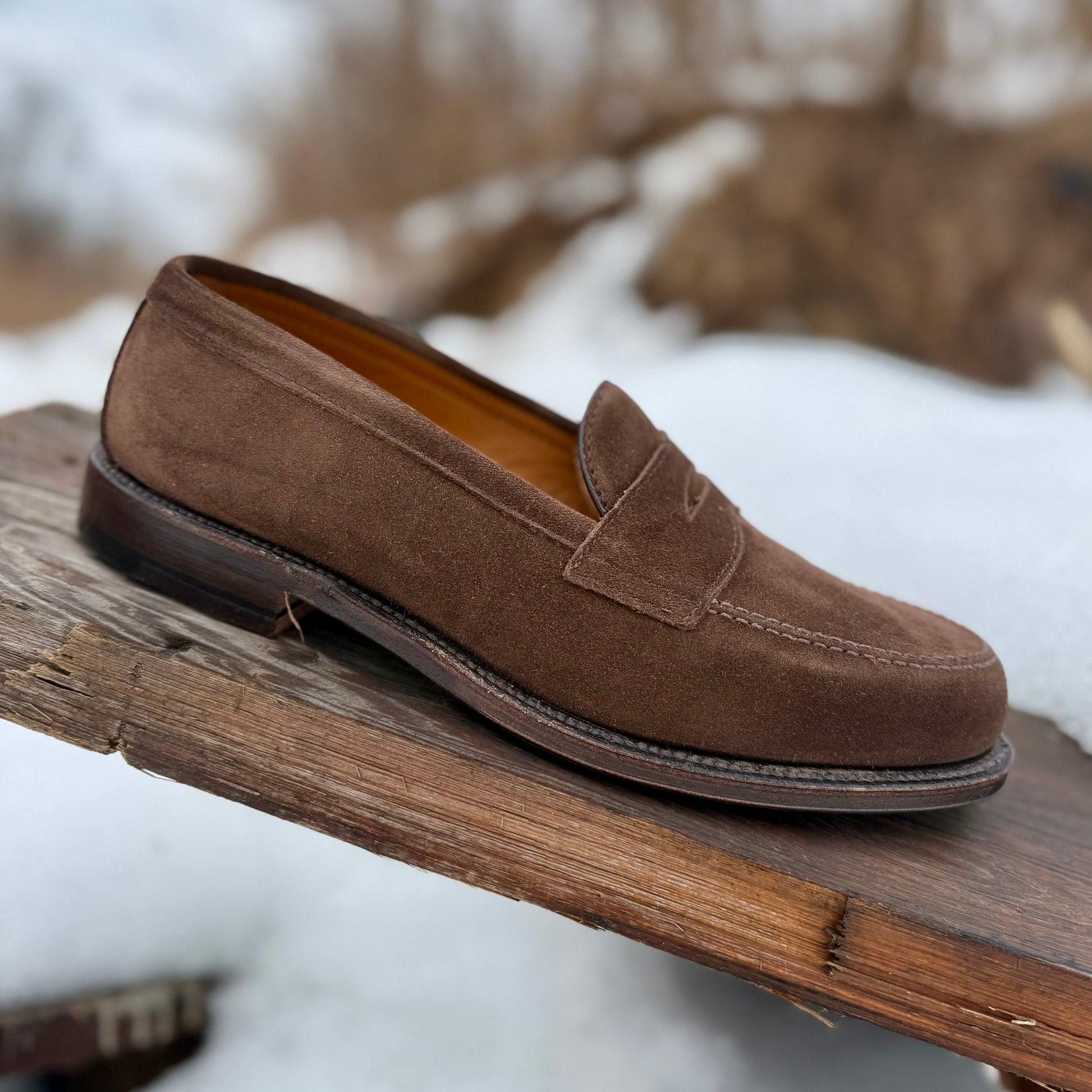 The Alden Madison 6175F Penny Loafer in Humus Suede, featuring a dark brown Flex Oiled Leather Sole, rests on a weathered wooden plank outdoors with snow and blurred natural scenery in the background.