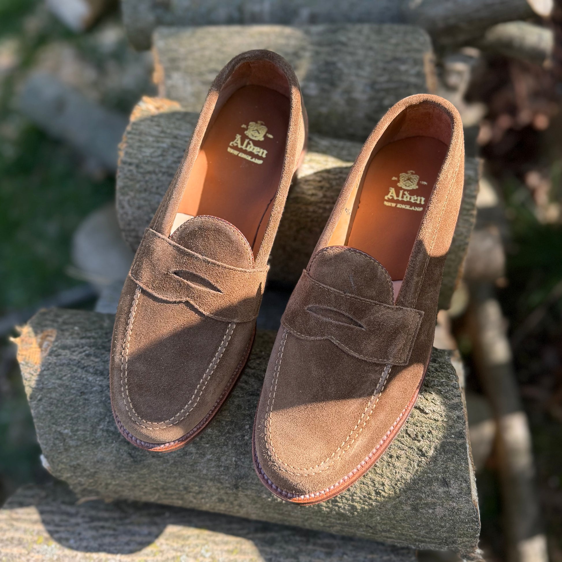 The Alden Madison 6173F Penny Loafer in Snuff Suede, featuring a Flex Leather Sole and crafted on the Van Last, is displayed outdoors on stacked logs in sunlight.