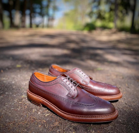The Alden Madison 976 - Long Wing Blucher in brown calfskin rests on a sunlit dirt path, its brogue detailing and double waterlock sole highlighted against blurred trees and greenery in the background.